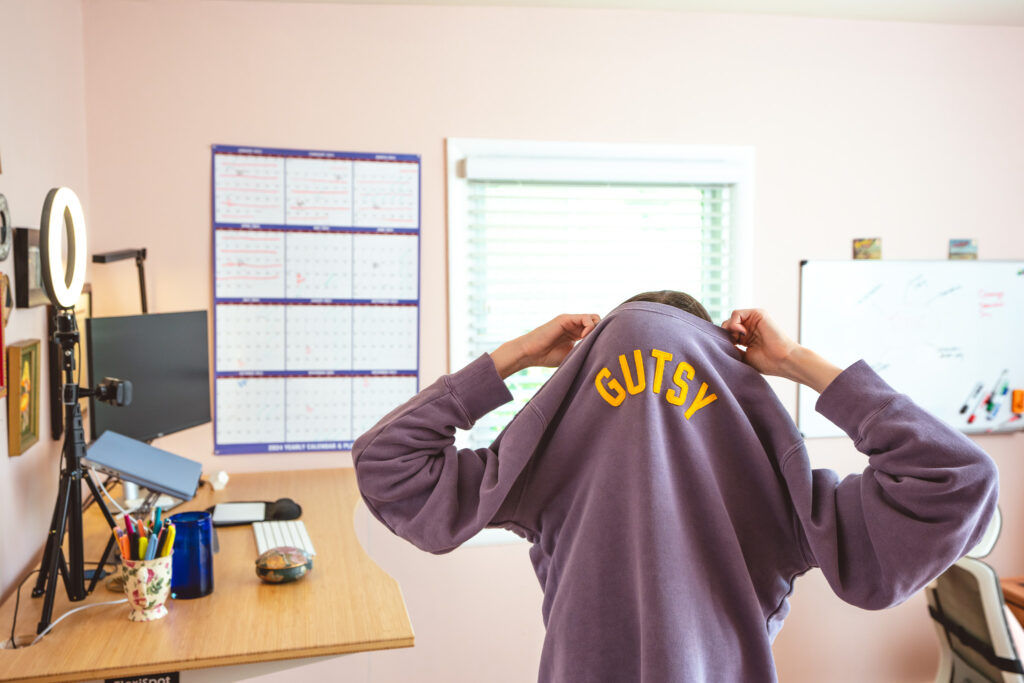 Copywriter Cassie Paton in her home office pulling a sweatshirt that says "GUTSY" over her head. She appears to be a little stuck.