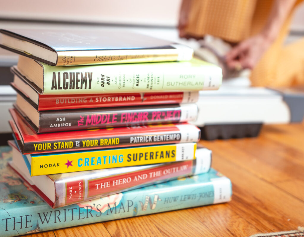 A stack of library books about branding, entrepreneurship, and writing. In the background, copywriter Cassie Paton puts her shoes on in preparation for firing up her walking pad.